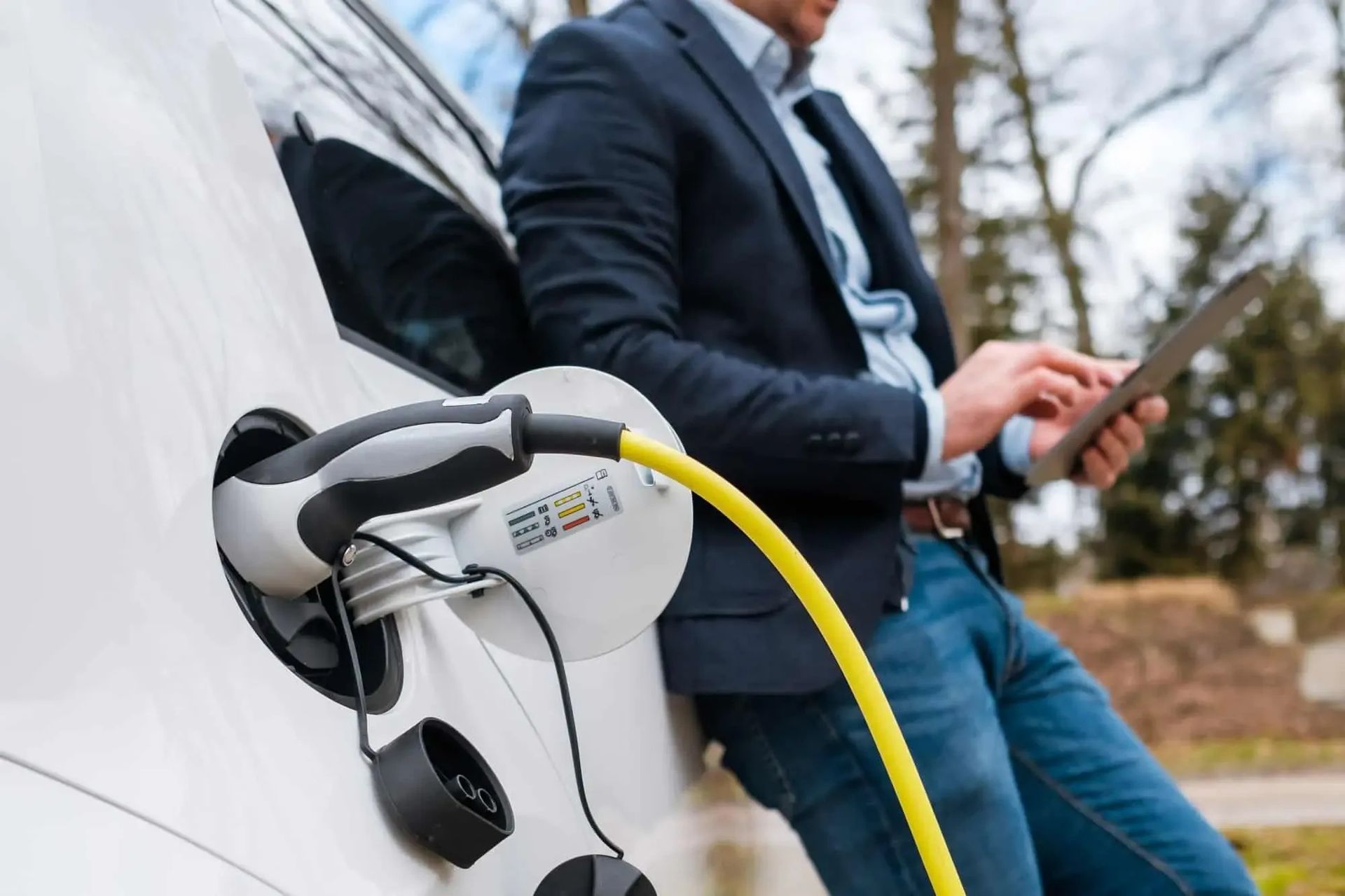 A person in a blazer and jeans uses a tablet while leaning against a white electric vehicle plugged into a yellow charger.