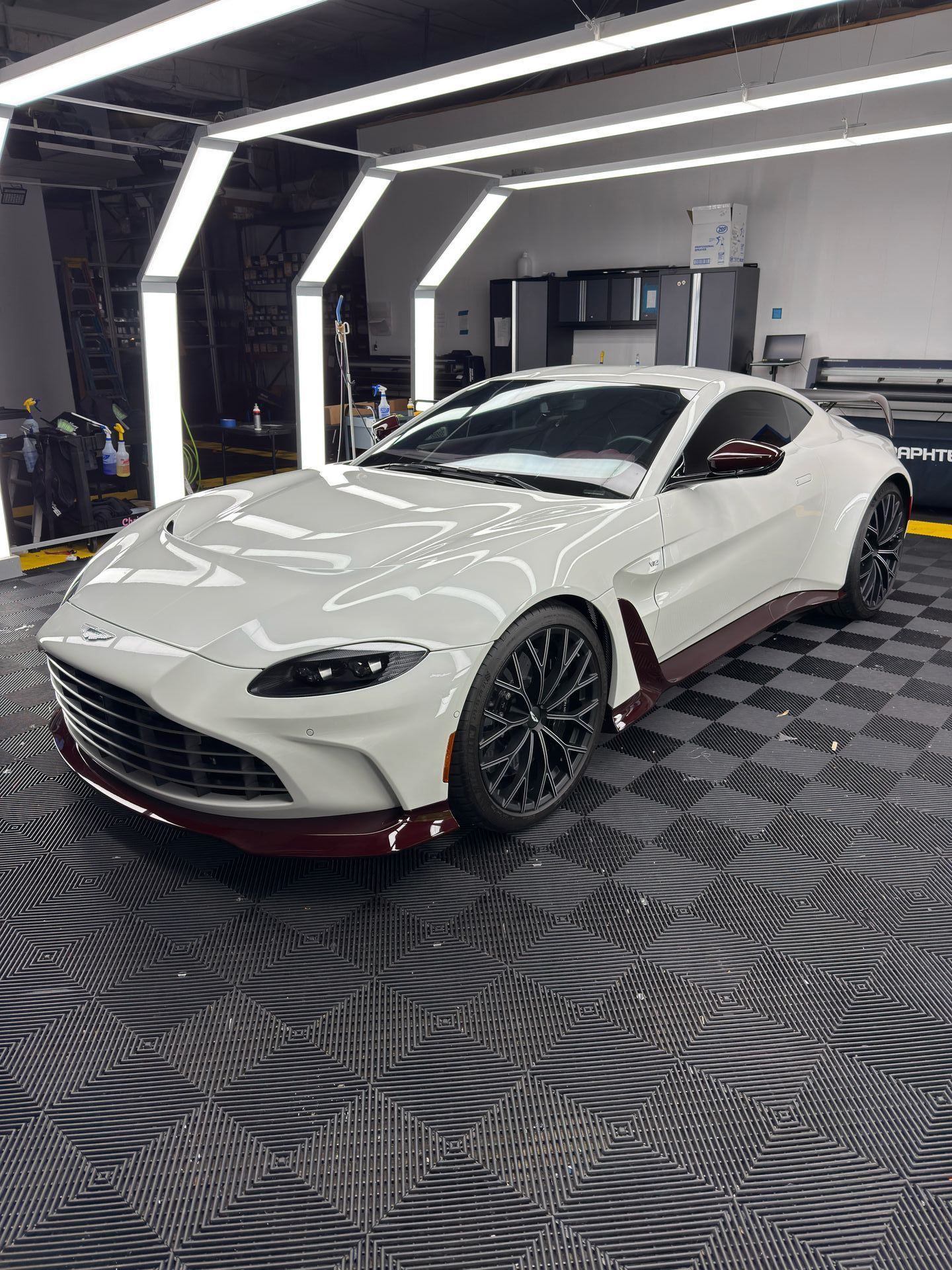 White Aston Martin sports car in a garage with black and gray checkered floor, under bright lights. 