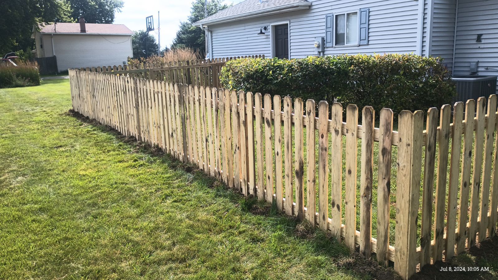 A newly installed natural wood picket fence runs alongside a house and a green lawn on a sunny day.
