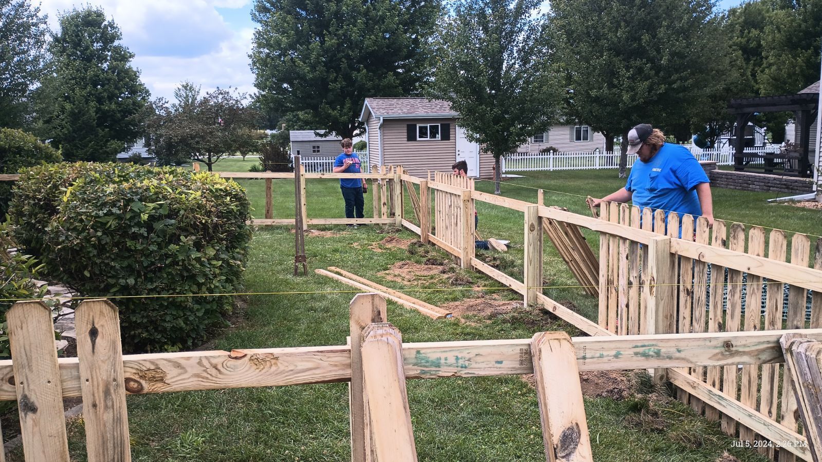 Two people in a yard construct a wooden picket fence around a grassy area near bushes and a small shed.