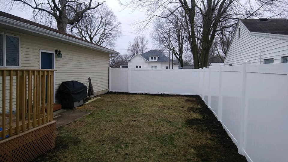 Backyard with white vinyl fence, wooden deck, and bare trees under overcast sky.