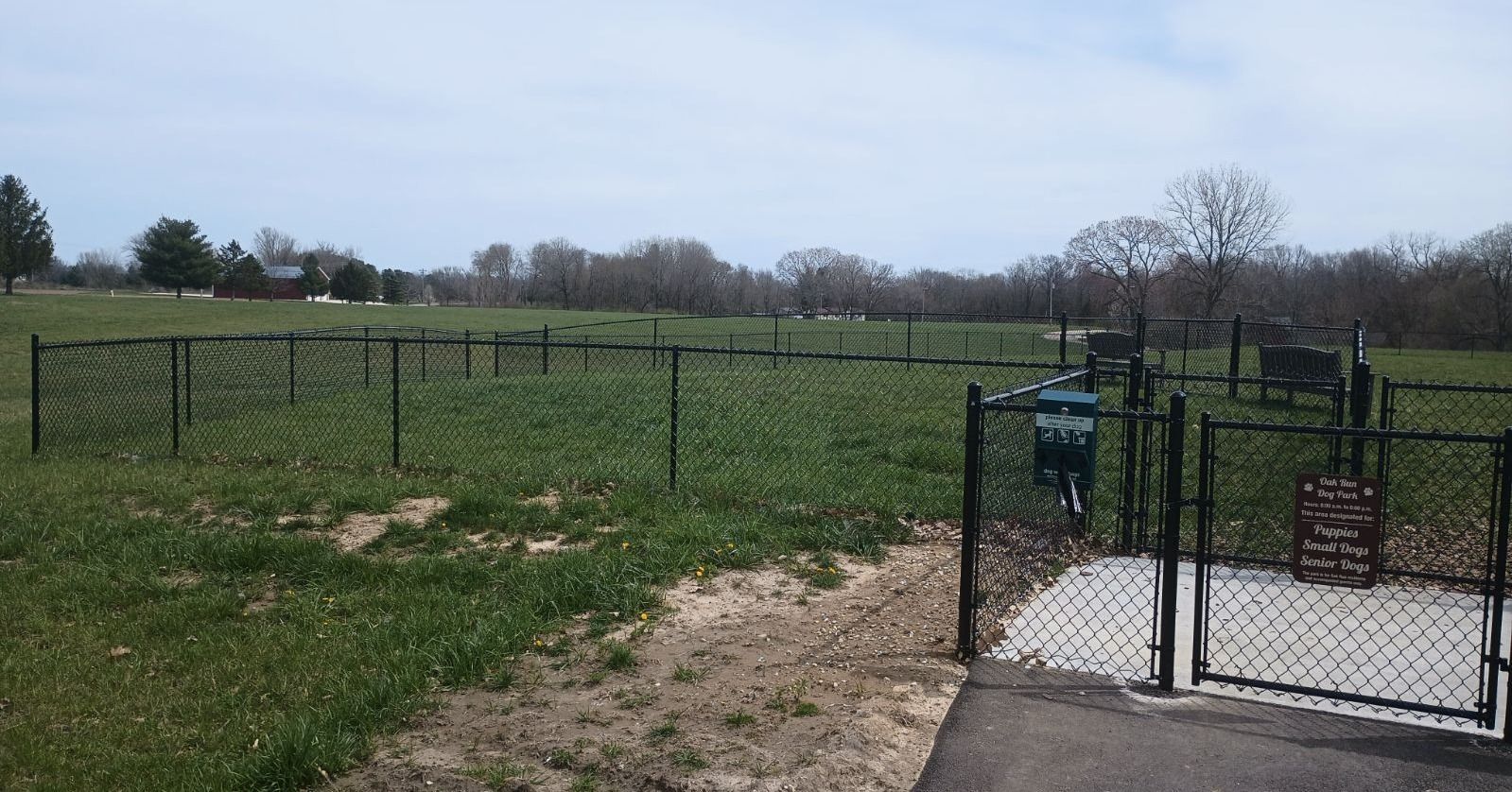 A fenced dog park area with a gated entrance on a grassy field under a cloudy sky.