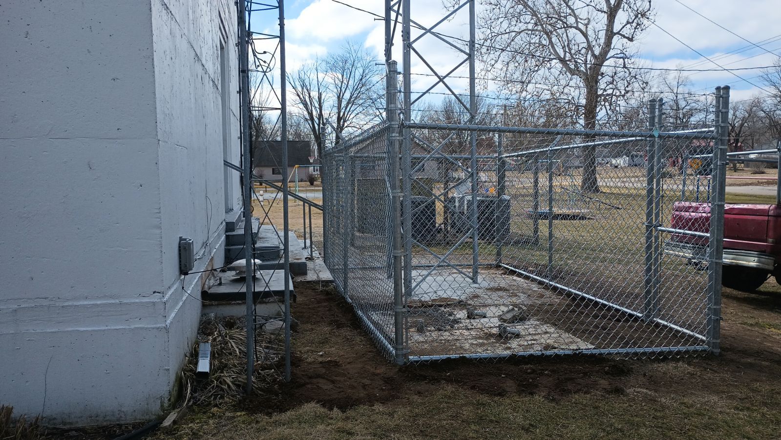 A chain-link fenced area surrounds the base of a metal communications tower next to a white building.