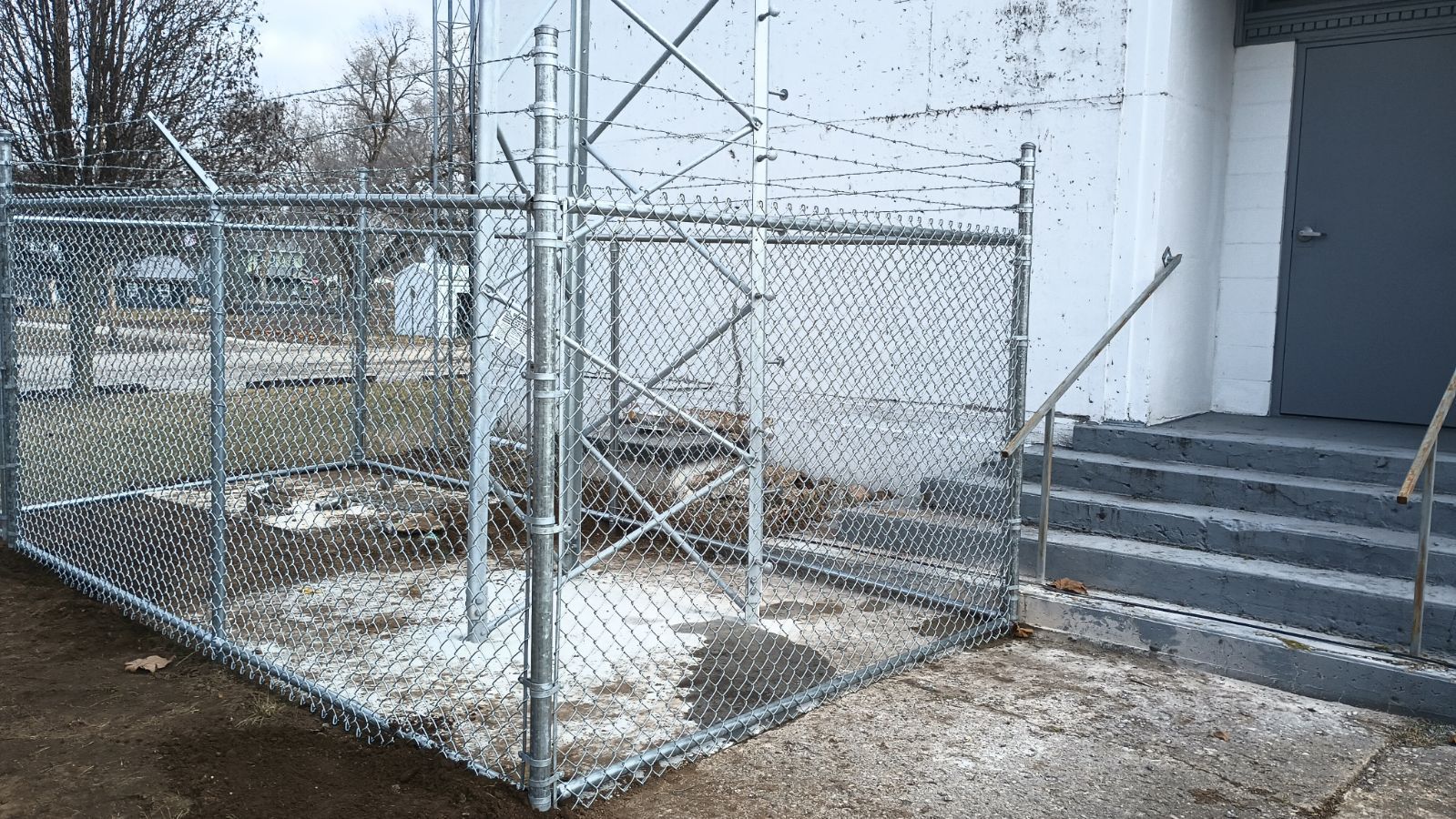 A chain-link fence topped with barbed wire surrounds a utility tower base next to a white building with concrete stairs.