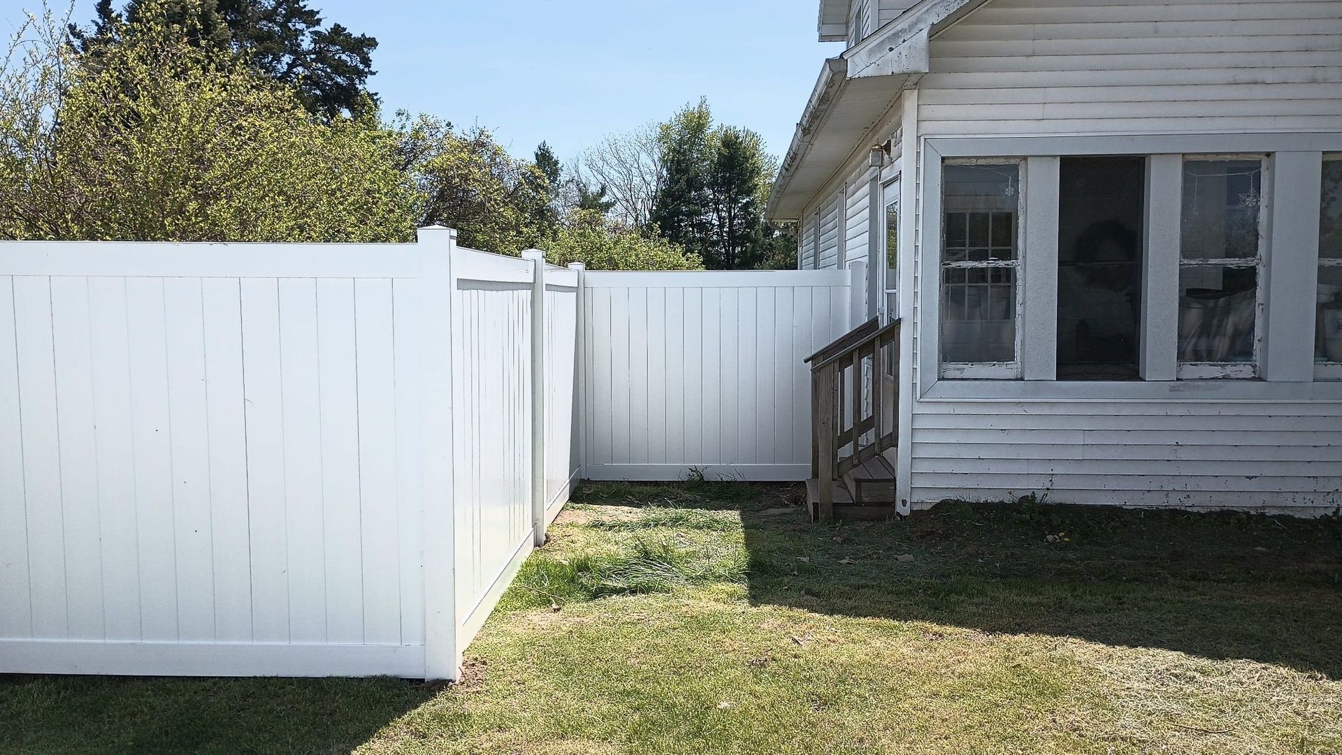 A white vinyl privacy fence meets the side of a white house with a porch step in a grassy yard under a clear blue sky.
