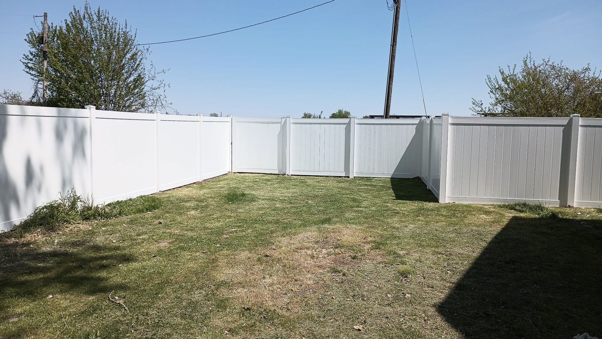 A small grassy backyard enclosed by a tall, white vinyl privacy fence under a clear blue sky.
