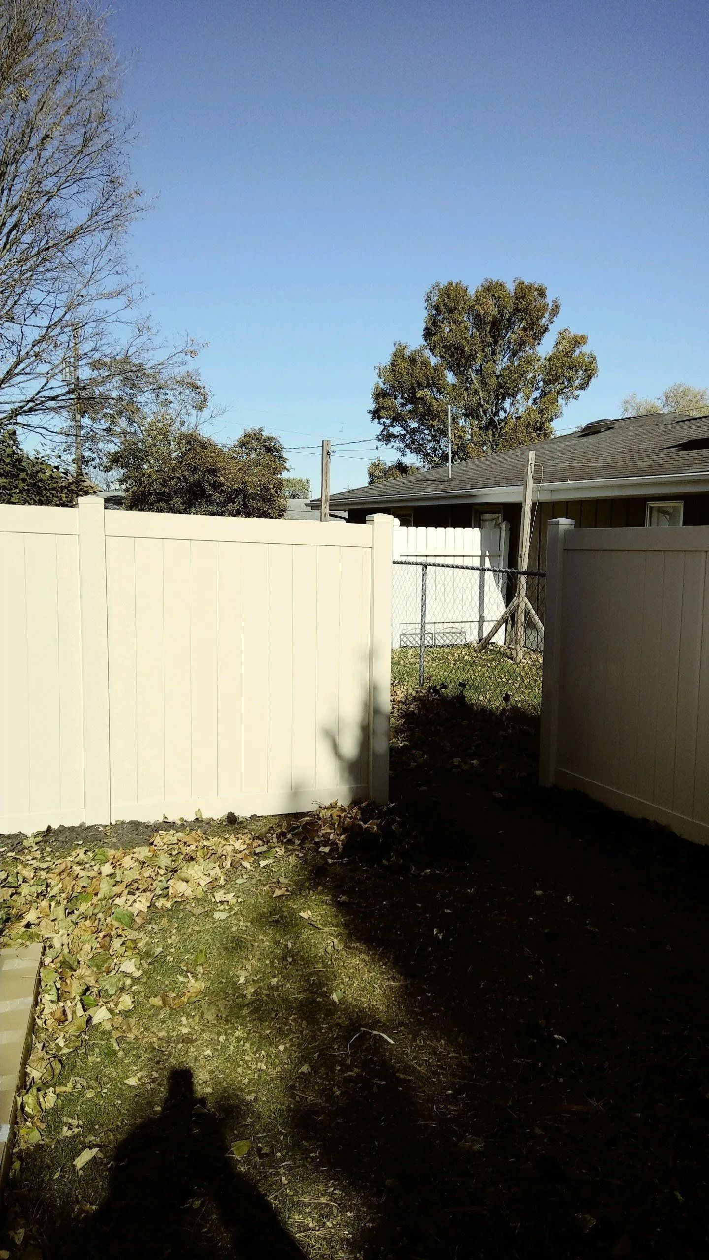 White fence bordering a grassy area and a dark pathway under a blue sky, with trees and a house in the background.