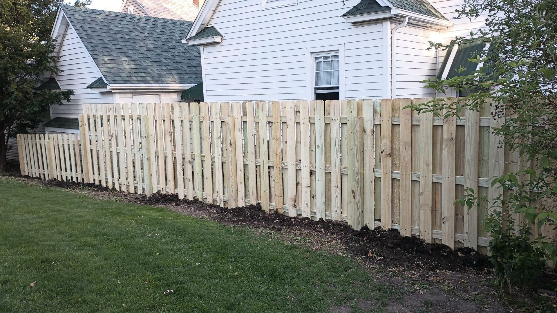 A newly installed, light-colored wooden picket fence stands in a grassy yard in front of a white house.
