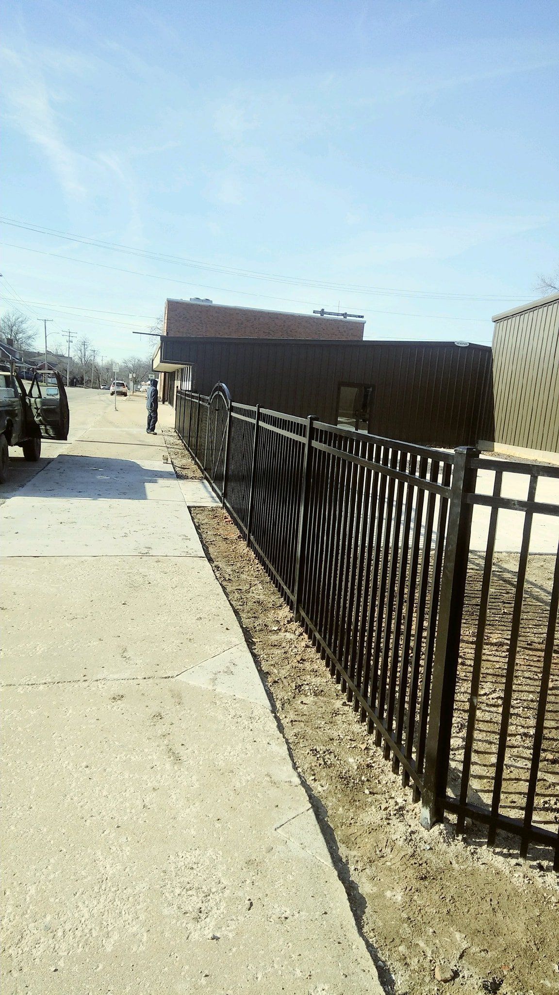 Black metal fence along a concrete sidewalk, with buildings in the background under a blue sky.