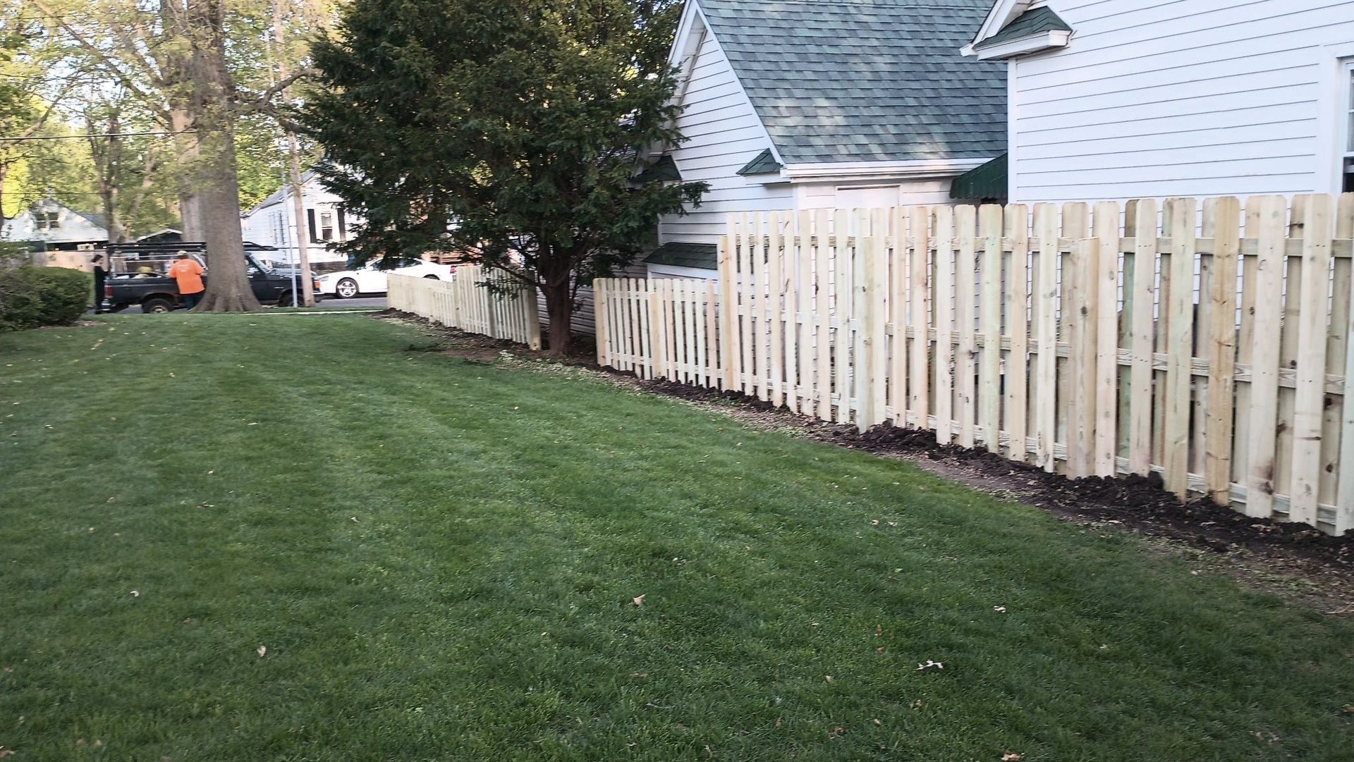 A newly installed light-colored wooden picket fence sits on the edge of a lush green lawn next to a white residential house.