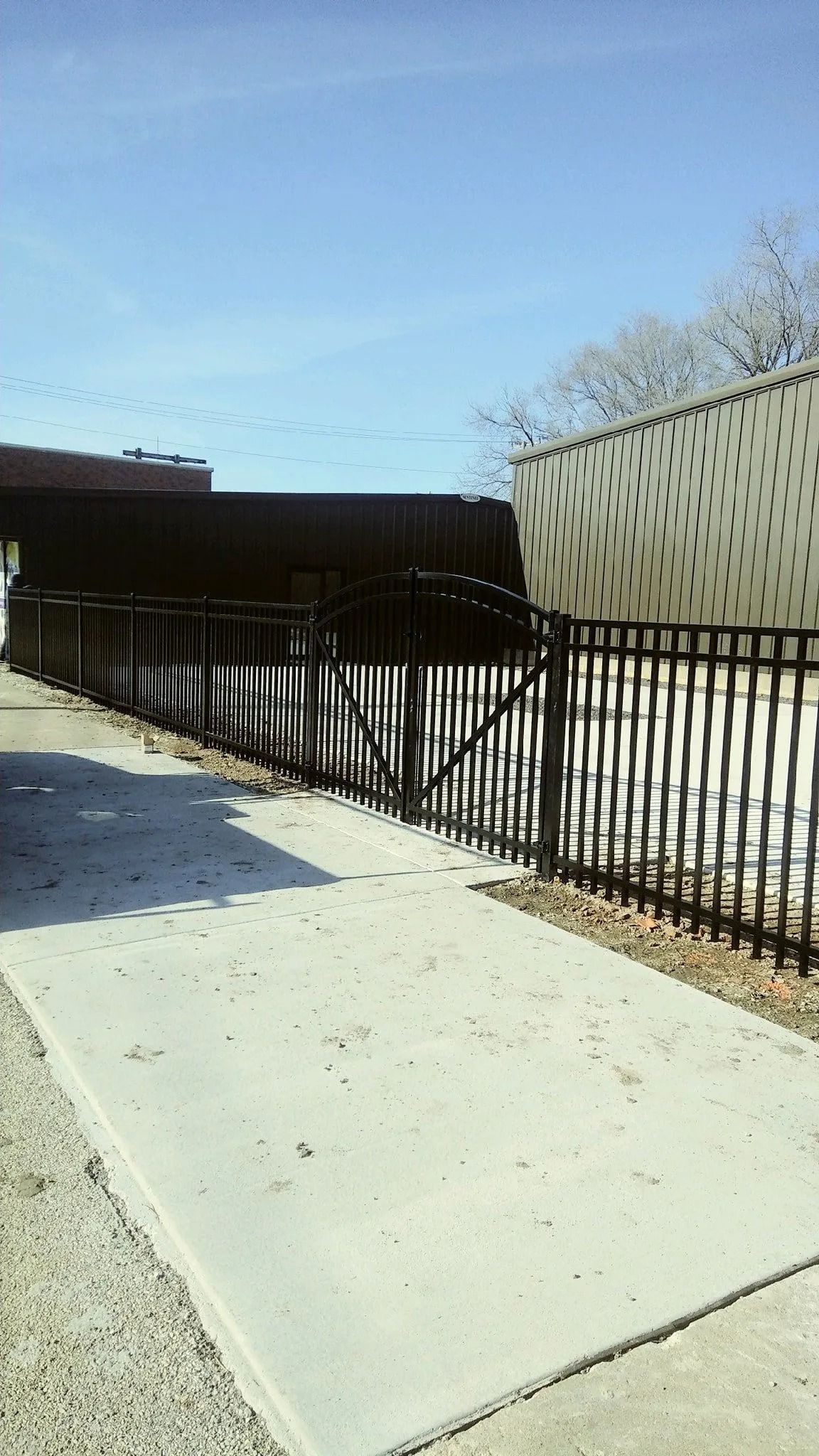 Sidewalk with a black fence, a metal building, and a bridge overhead against a blue sky.