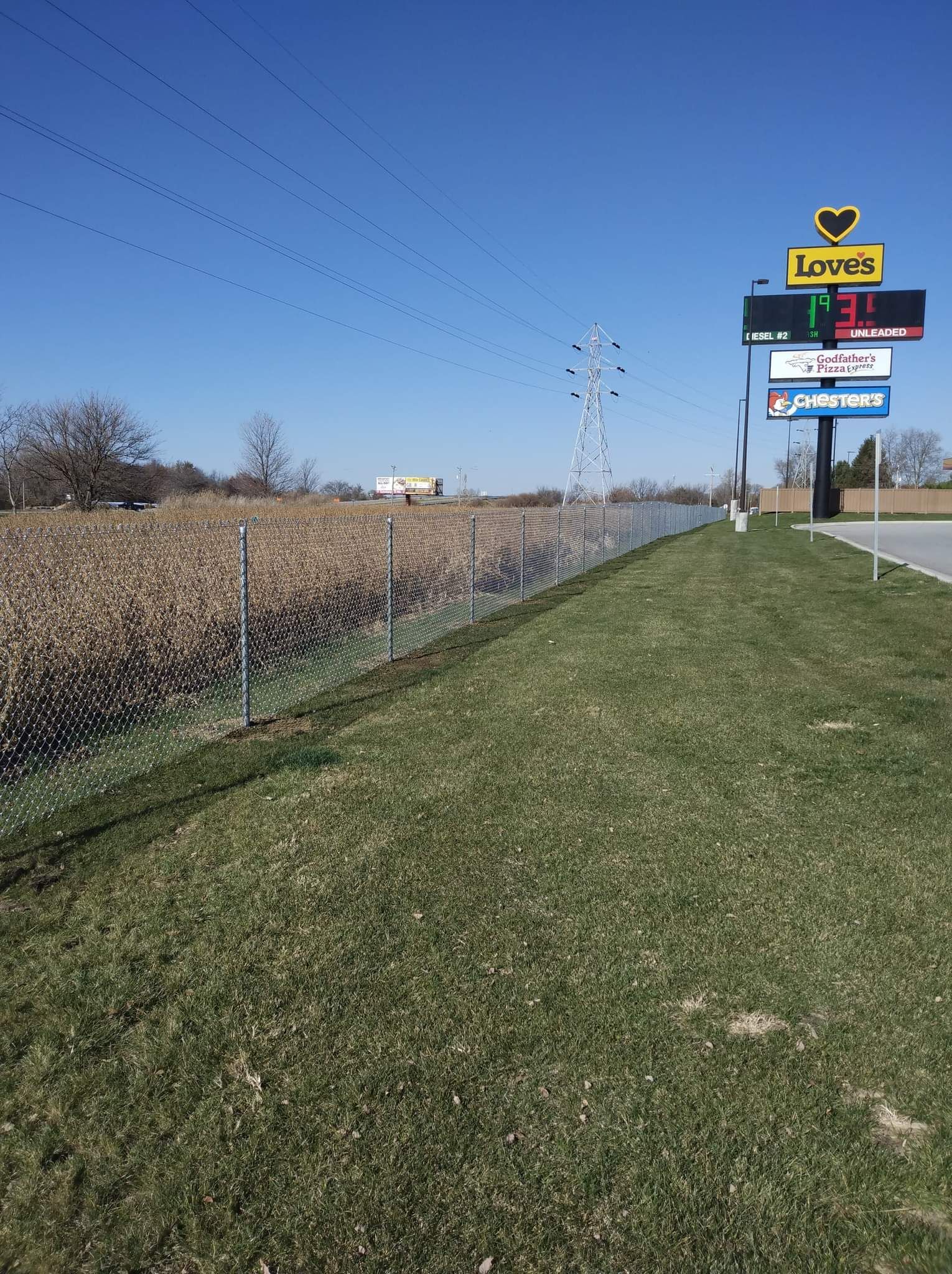 A chain-link fence lines a grassy lot next to a Lowe’s sign displaying a fuel price under a clear blue sky.