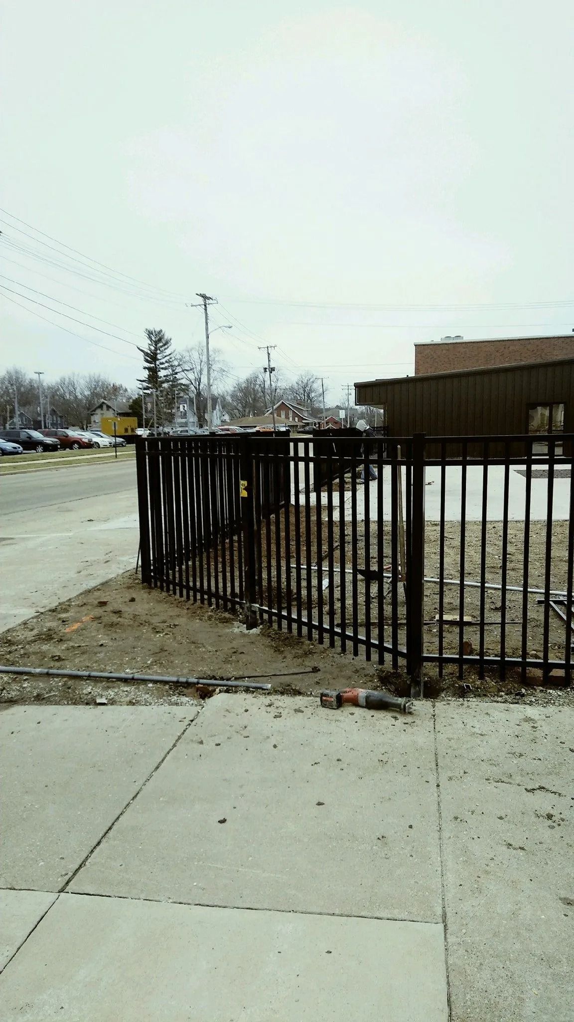 Black metal fence bordering a dirt lot, sidewalk in the foreground, cloudy sky.