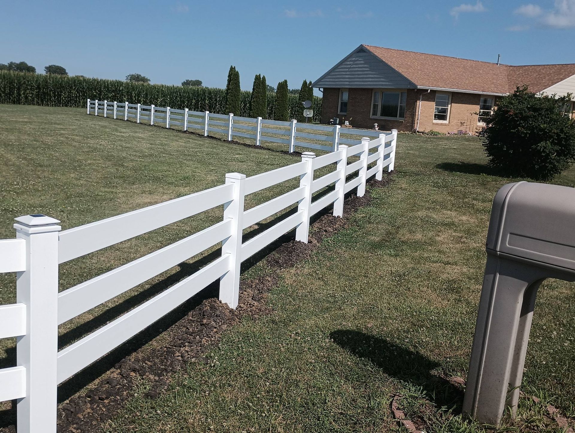 A white vinyl fence borders a grassy yard in front of a tan suburban house under a clear blue sky.