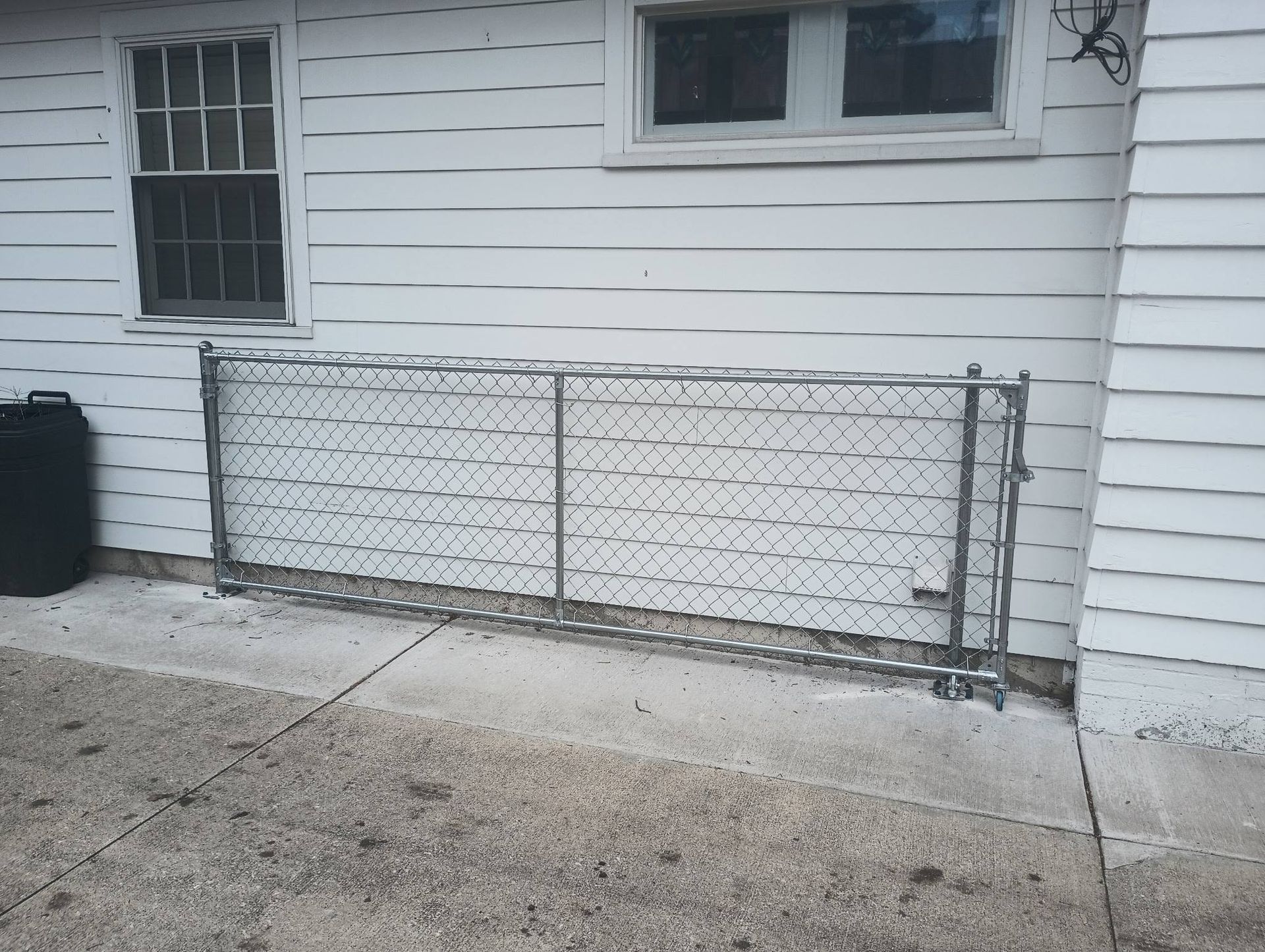 A section of chain-link fence leans against the white-sided exterior wall of a building on a concrete patio.