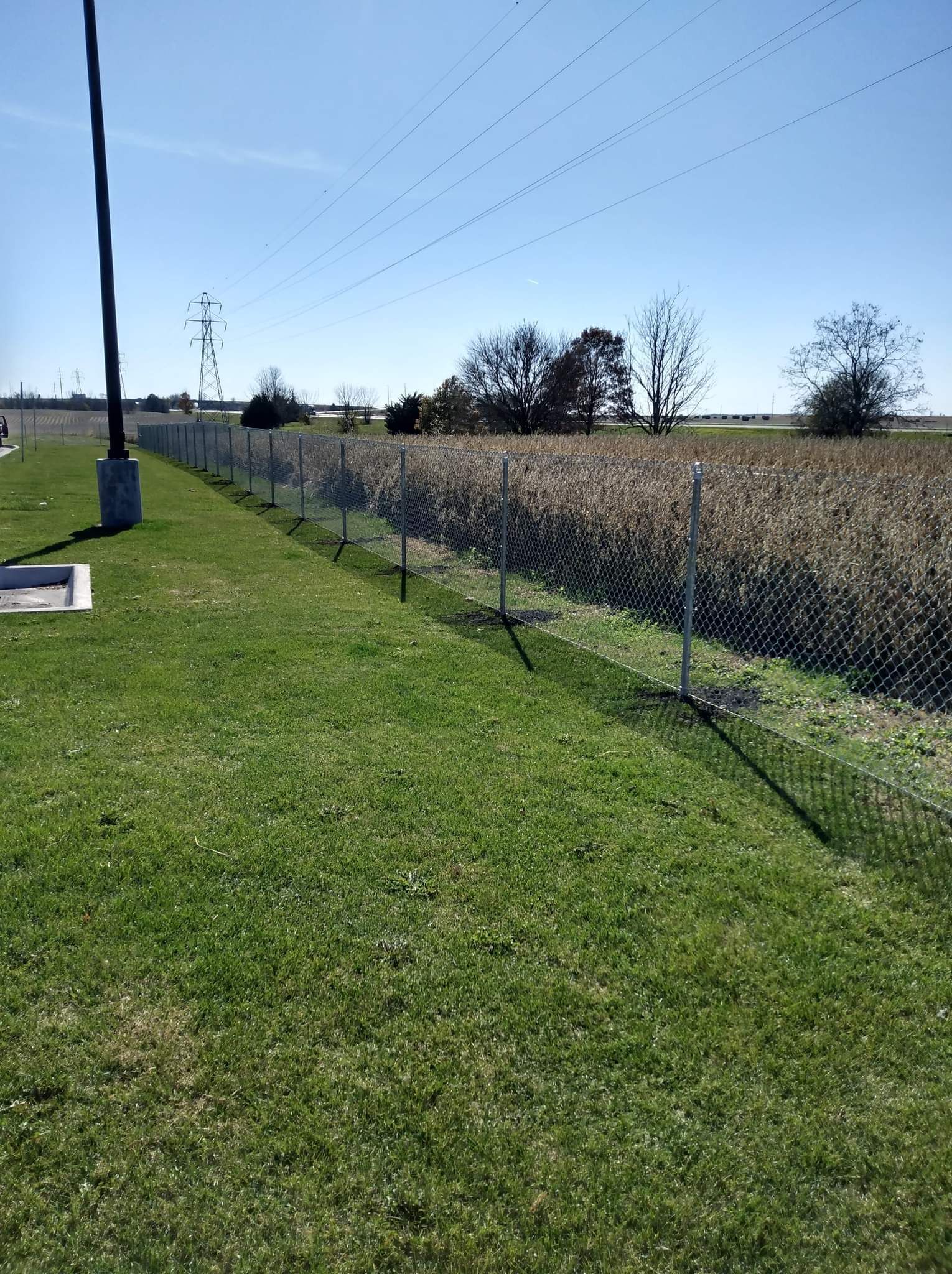 A chain-link fence divides a grassy foreground from a large field of harvested crops under a clear blue sky.