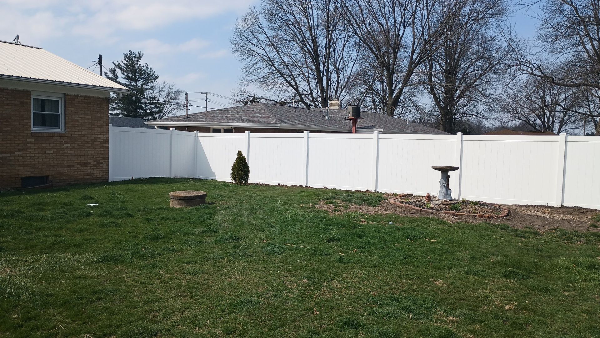 A backyard featuring a brick house, a white vinyl fence, a small evergreen bush, a birdbath, and a patch of grass.