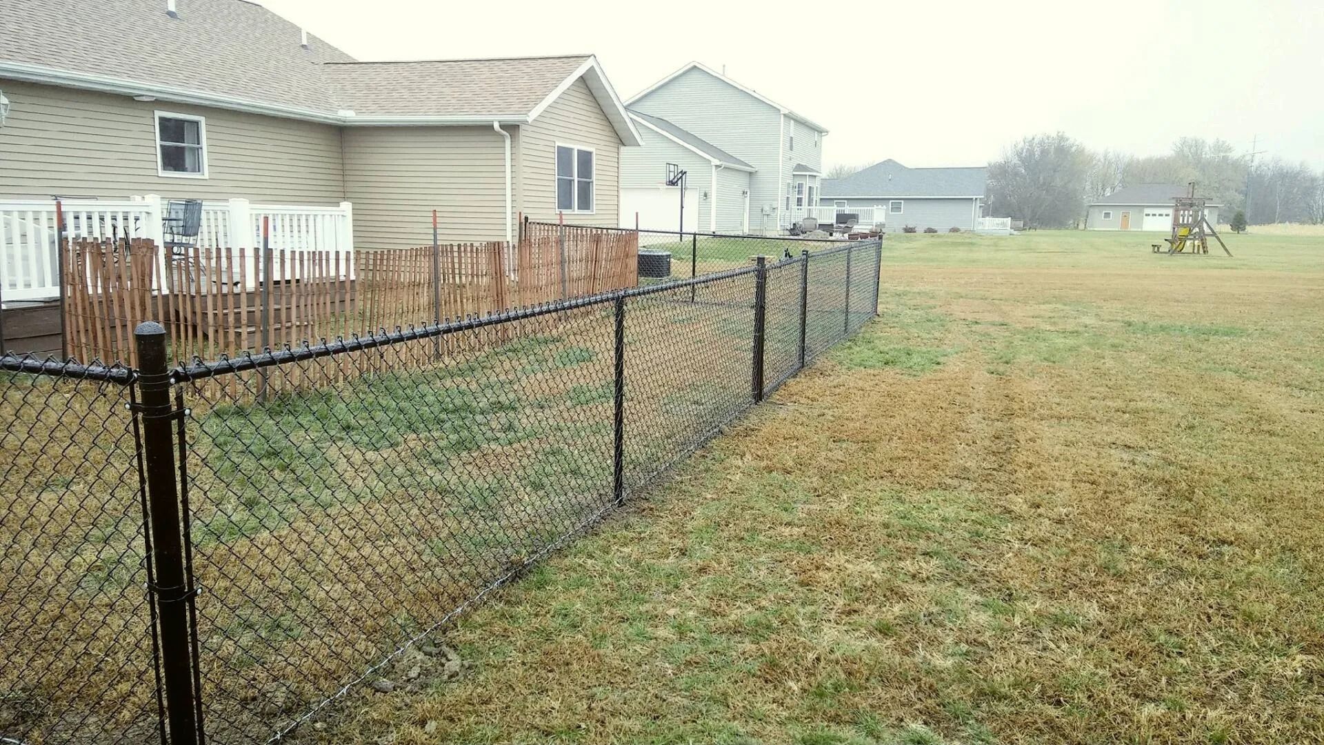 Black chain-link fence bordering a grassy yard, beige houses in the background under an overcast sky.