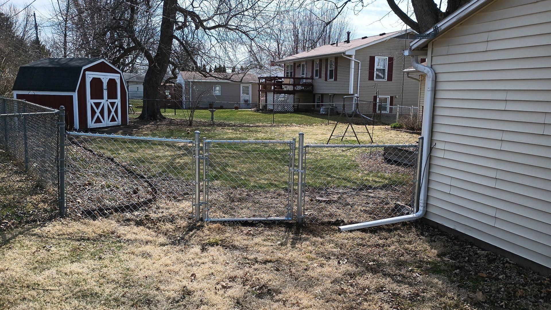 A chain-link fence with a center gate separates two backyards, one featuring a small red shed and the other a house.