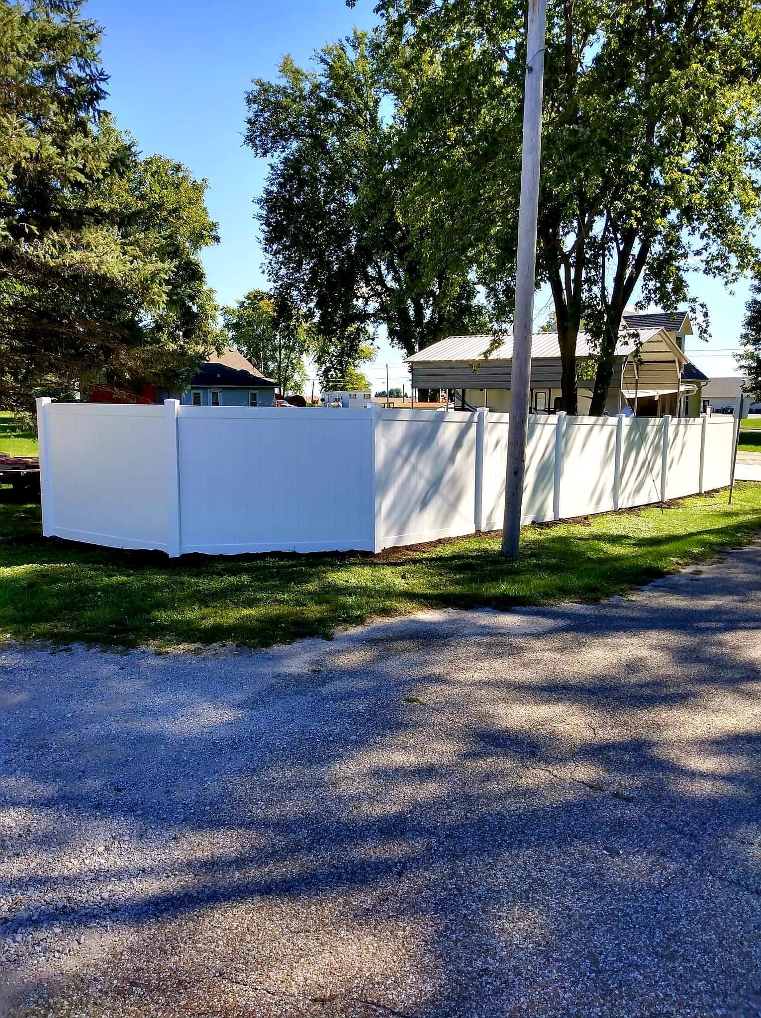 A white vinyl privacy fence encloses a backyard with trees and a house in the background under a clear blue sky.