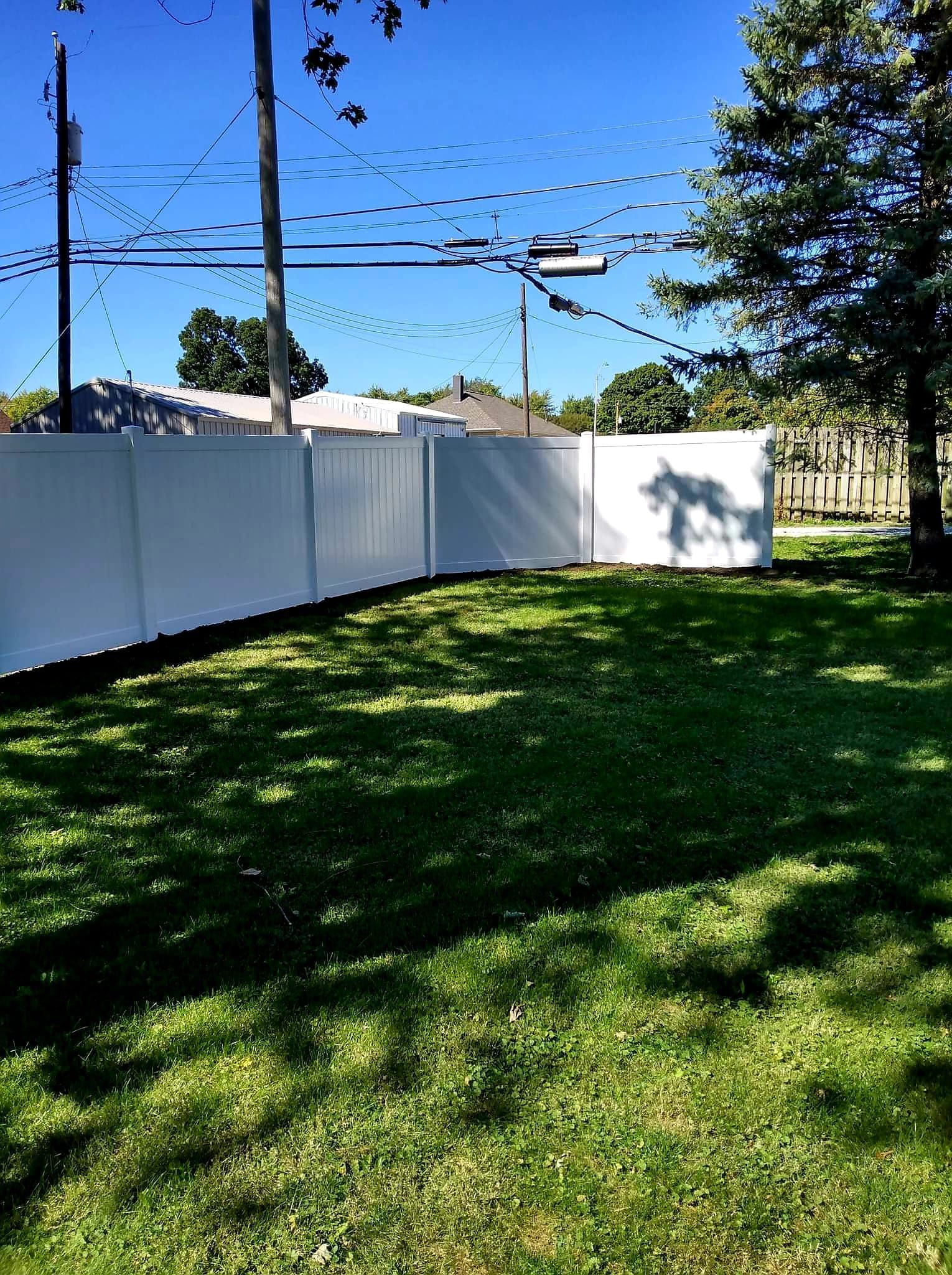 A white vinyl privacy fence segments a green backyard on a sunny day with shadows cast on the grass.