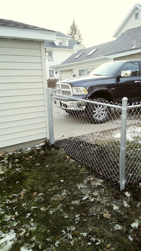 Black pickup truck behind a chain-link fence, next to a beige-sided building. Grassy yard with patches of snow.