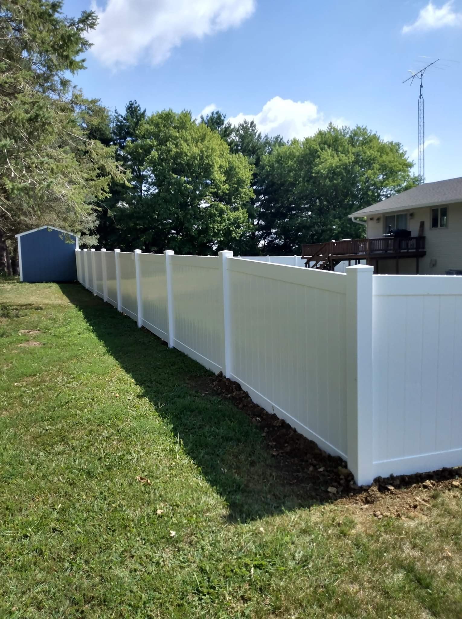 A white vinyl privacy fence lines a backyard, separating the lawn from a house and trees under a sunny blue sky.