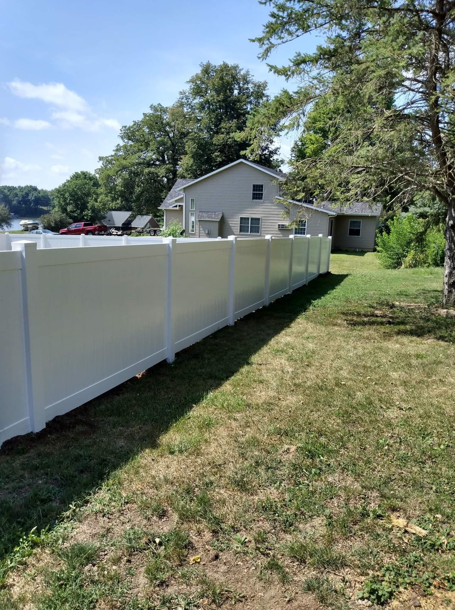 A long white vinyl privacy fence lines a grassy yard on a sunny day with a house in the background.