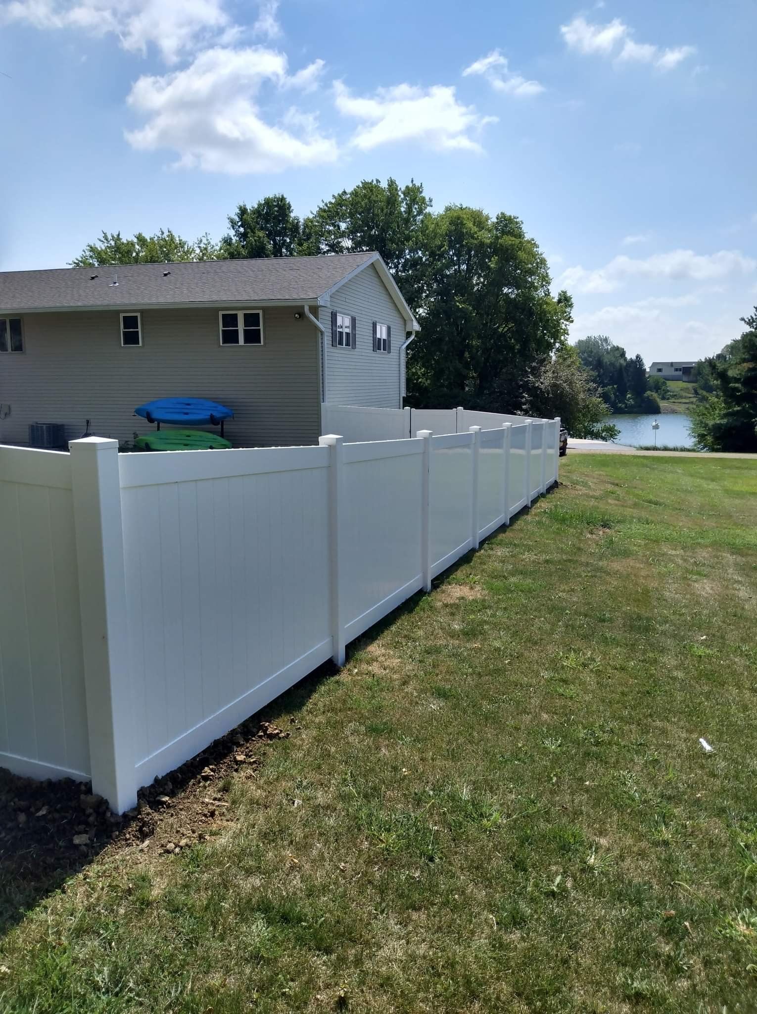 A tall, white vinyl privacy fence lines the grassy edge of a backyard, with a house and a distant lake in the background.