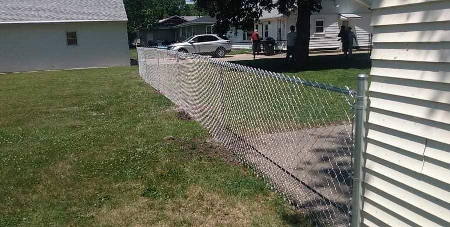 Chain-link fence borders a grassy yard, beside a paved pathway. A white house is visible.