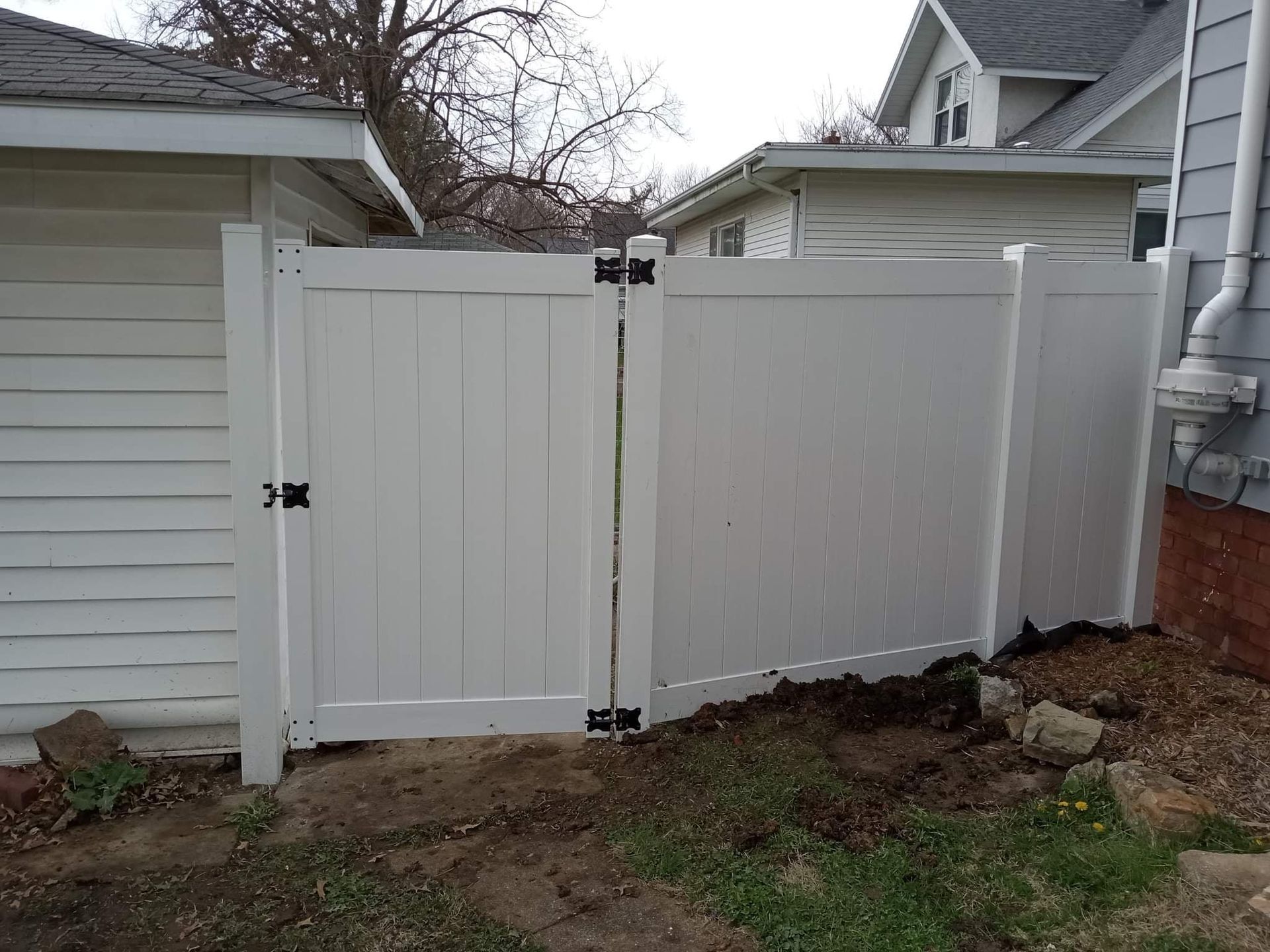 A white vinyl gate and fence installed between two house structures, situated over a patch of dirt and sparse grass.