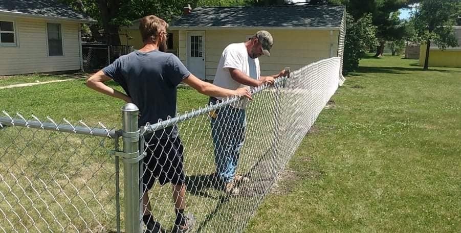 Two people installing a chain-link fence in a grassy yard on a sunny day.