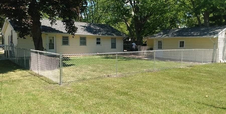 A chain-link fence encloses a green lawn with two small, light yellow houses in the background on a sunny day.