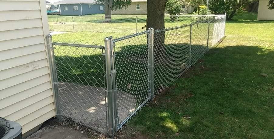 Chain-link fence section with gate, attached to a house, enclosing a small yard area, with grass and a tree.
