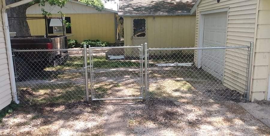 Chain link fence with gate, between two yellow buildings and a truck.