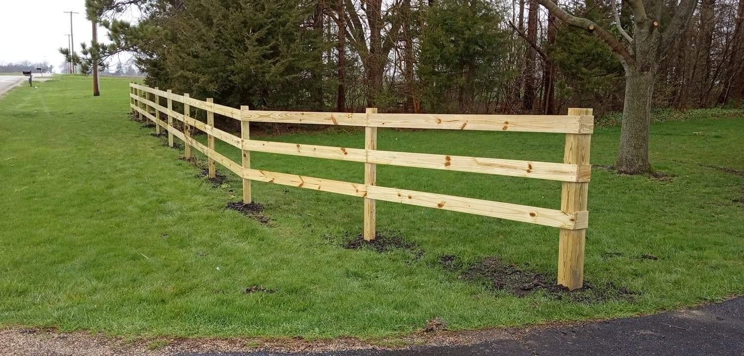 A three-rail wooden fence curves along a green grassy lawn beside trees and a gravel road.