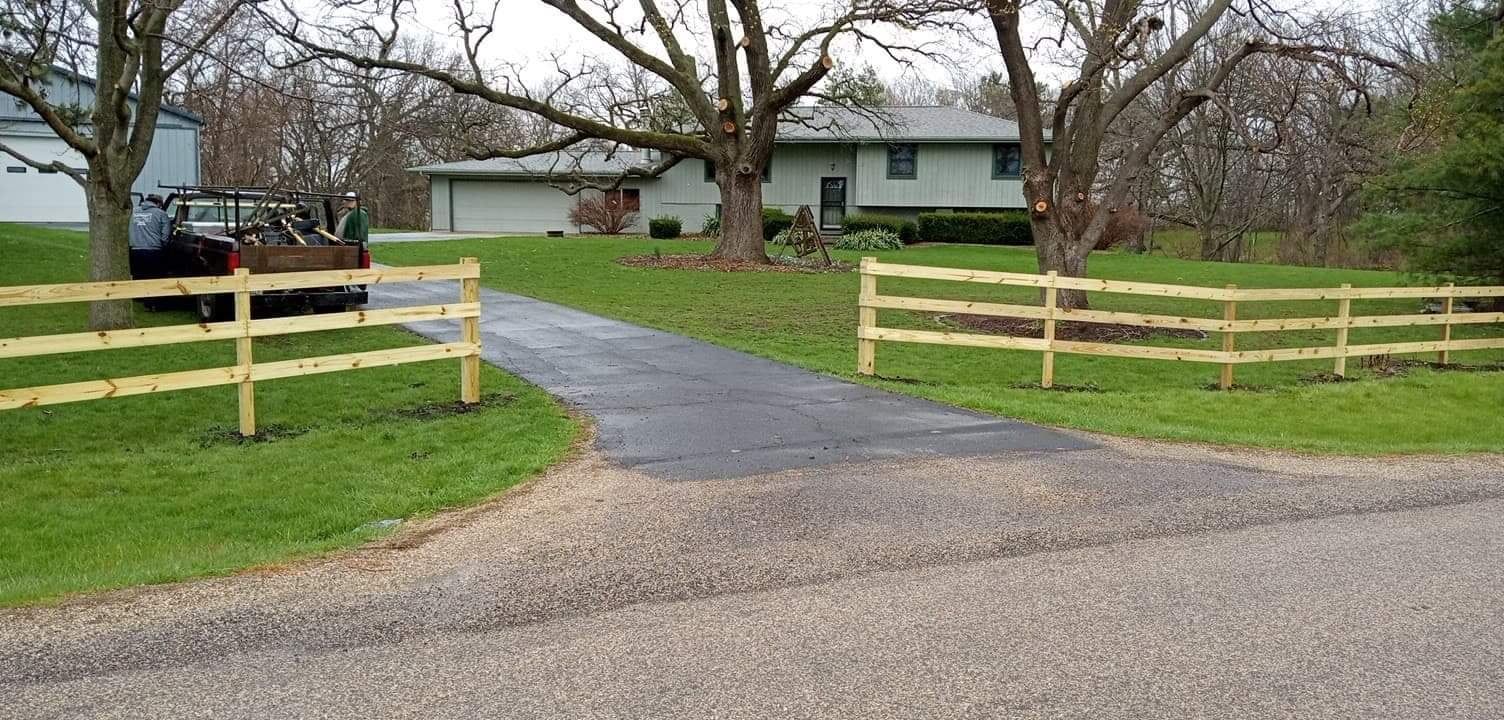 A split-rail wooden fence lines both sides of a paved driveway leading to a light green house on a grassy lawn.