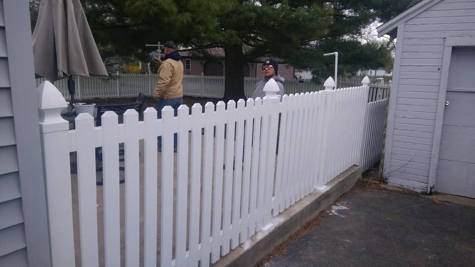 White picket fence next to a gray building and garage. Two people stand behind the fence.