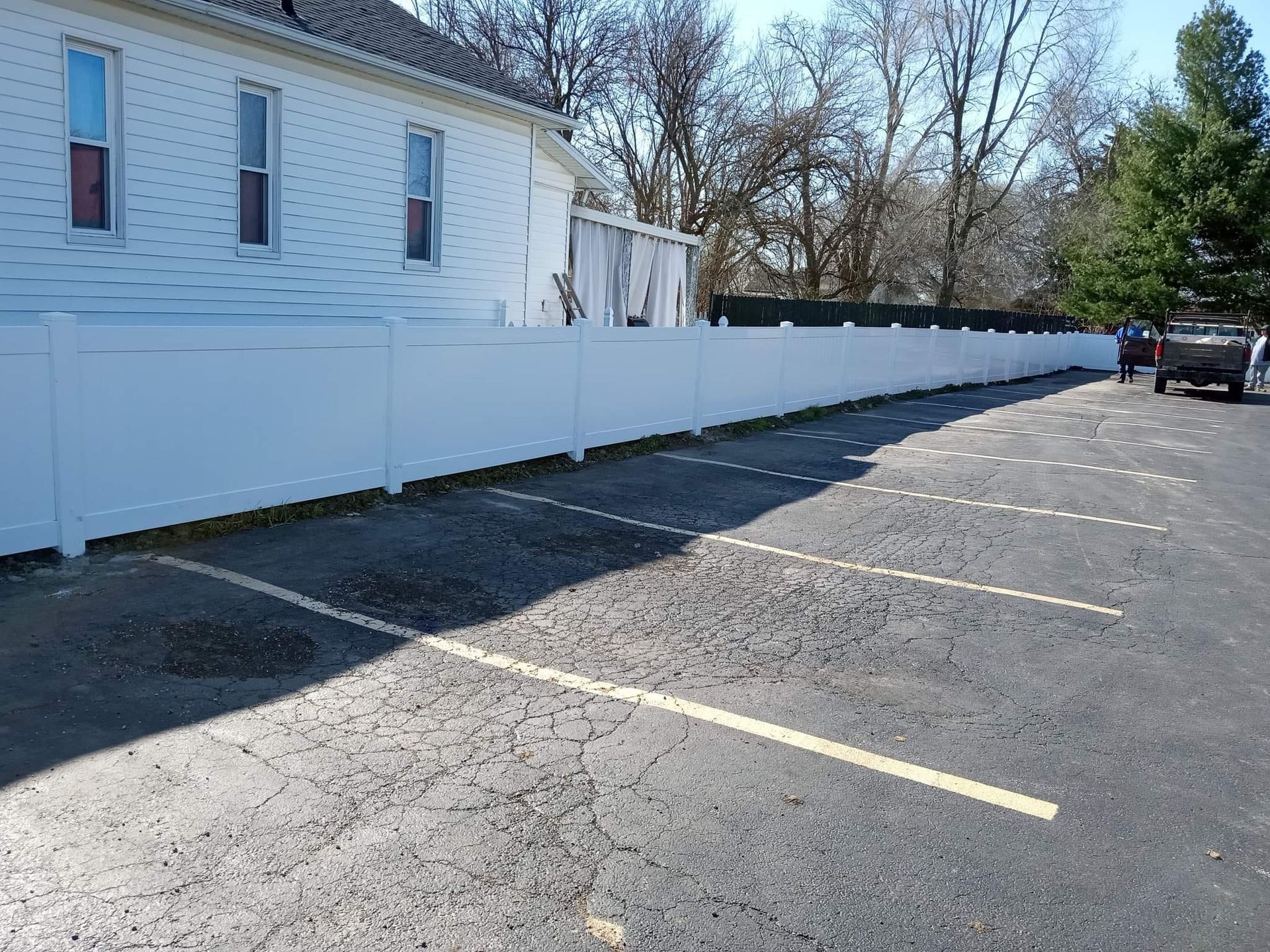 White vinyl fence bordering an asphalt parking lot with painted parking stalls, next to a white building.