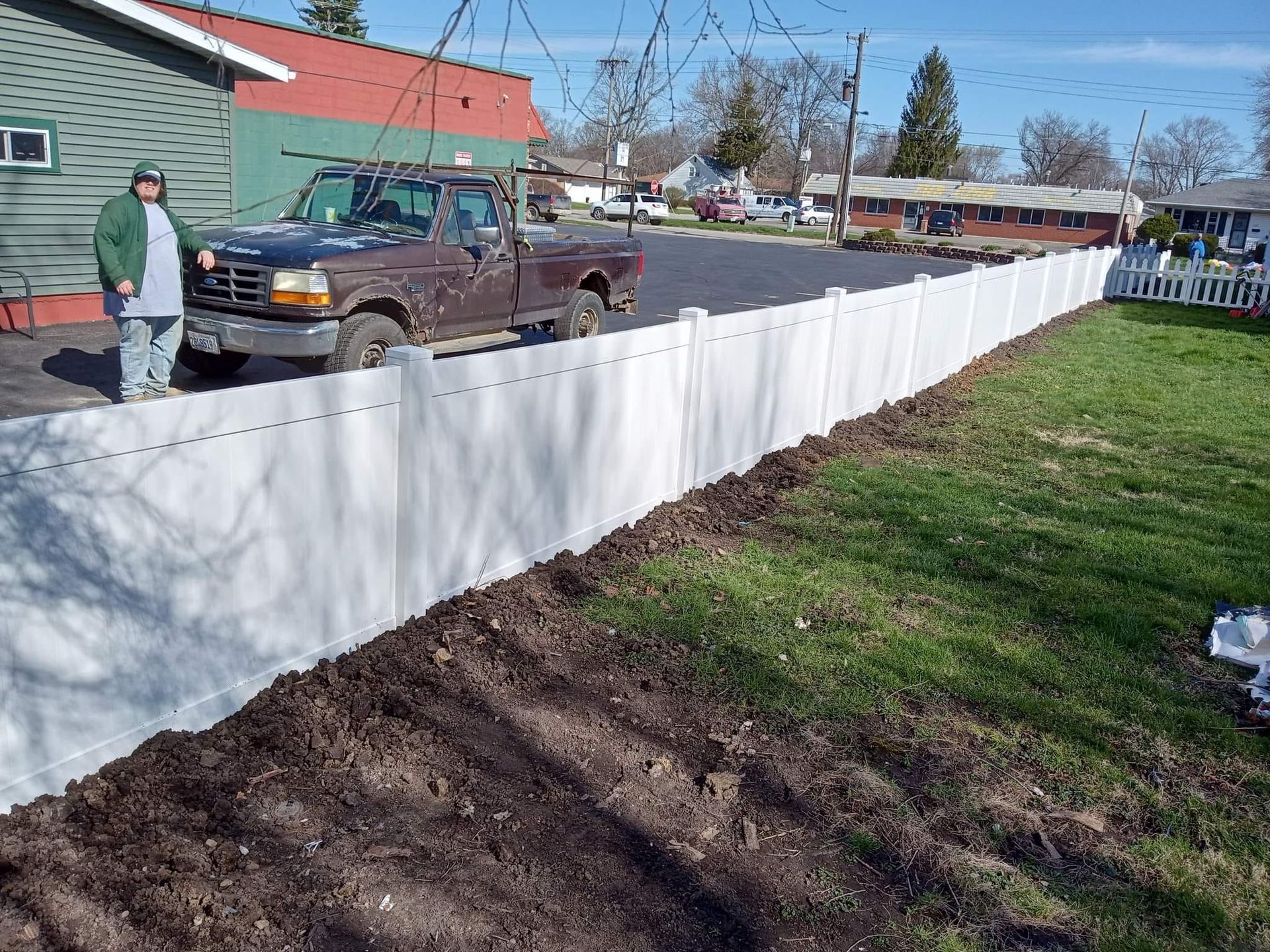A person in a green jacket stands beside a dark pickup truck next to a newly installed long white vinyl fence.