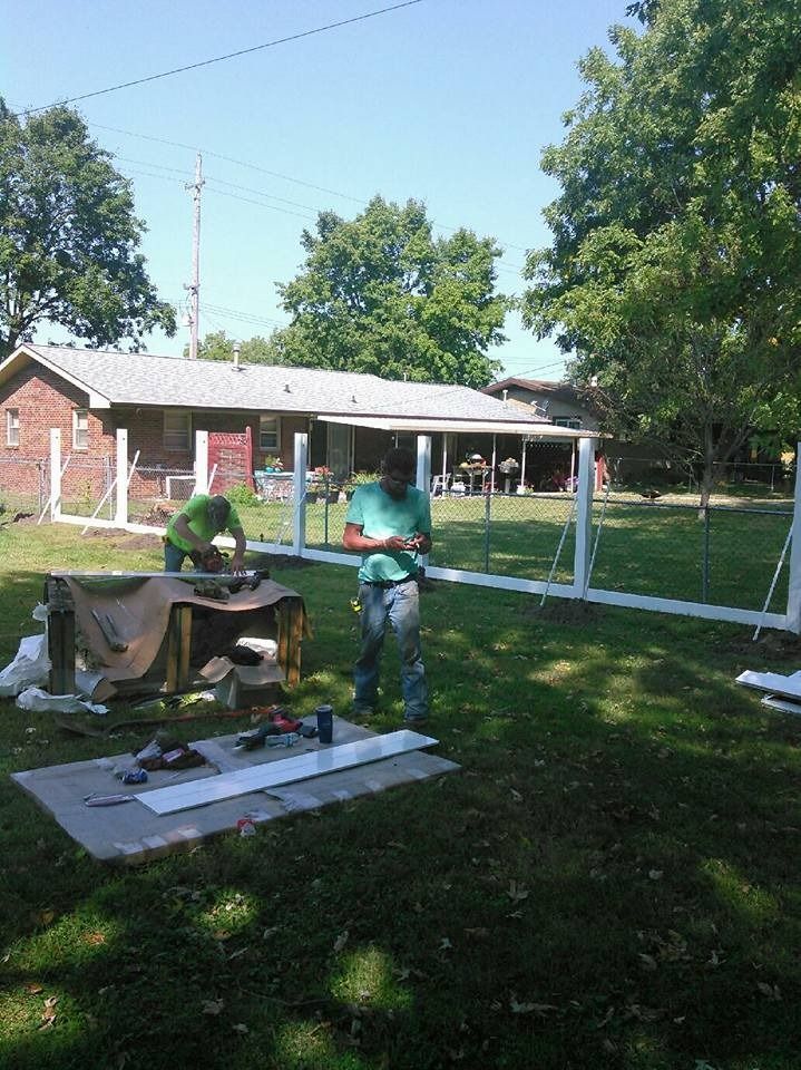 Two people building a white fence in a backyard near a house.