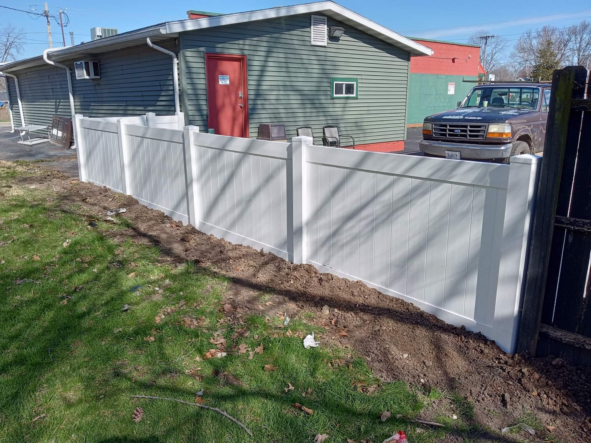 A white vinyl privacy fence stands in a grassy yard in front of a green building and a parked pickup truck.