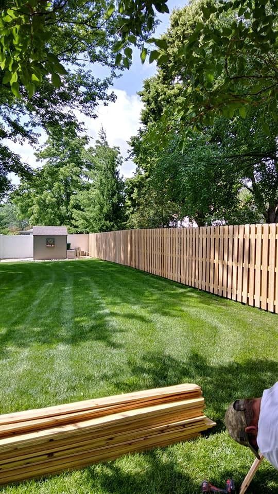 Lush green backyard with a new wooden fence, shed, and stacks of lumber on the grass under a blue sky.