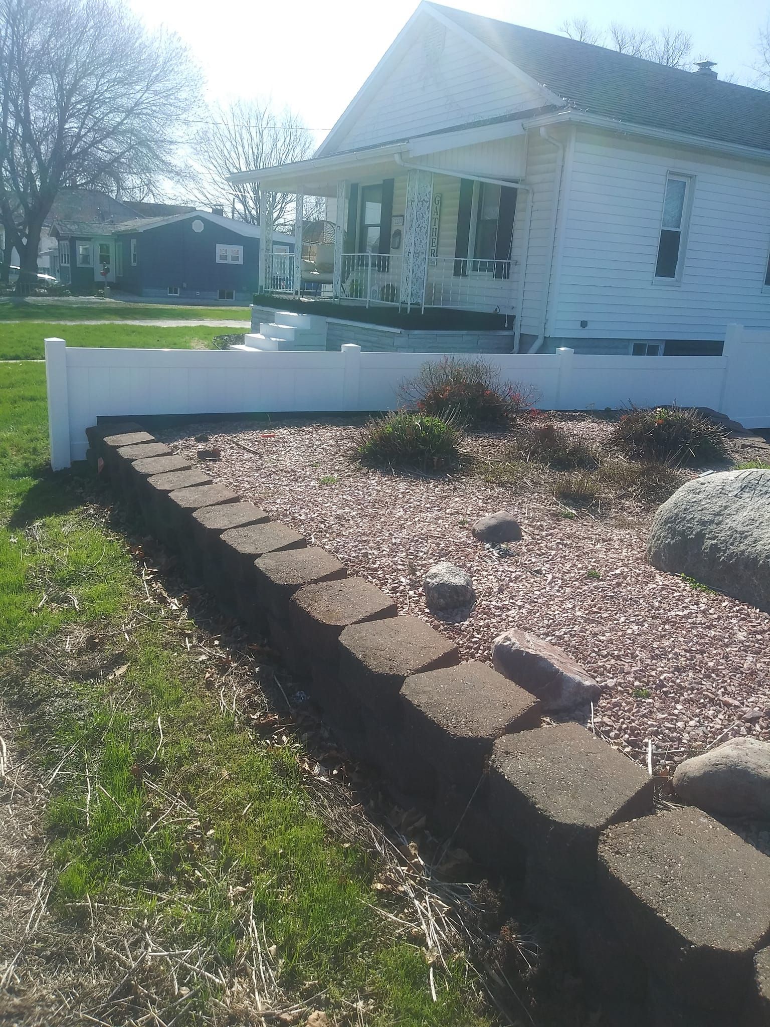 A low stone retaining wall borders a yard of decorative gravel and shrubs in front of a white house with a porch.