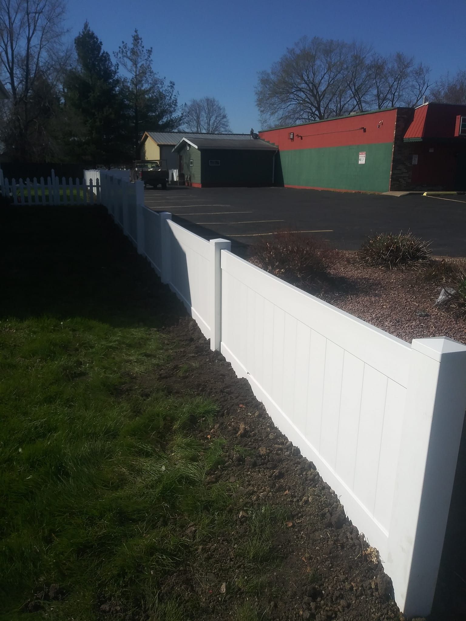 A white vinyl picket fence stands along the edge of a grassy lawn, with a paved lot and building in the background.
