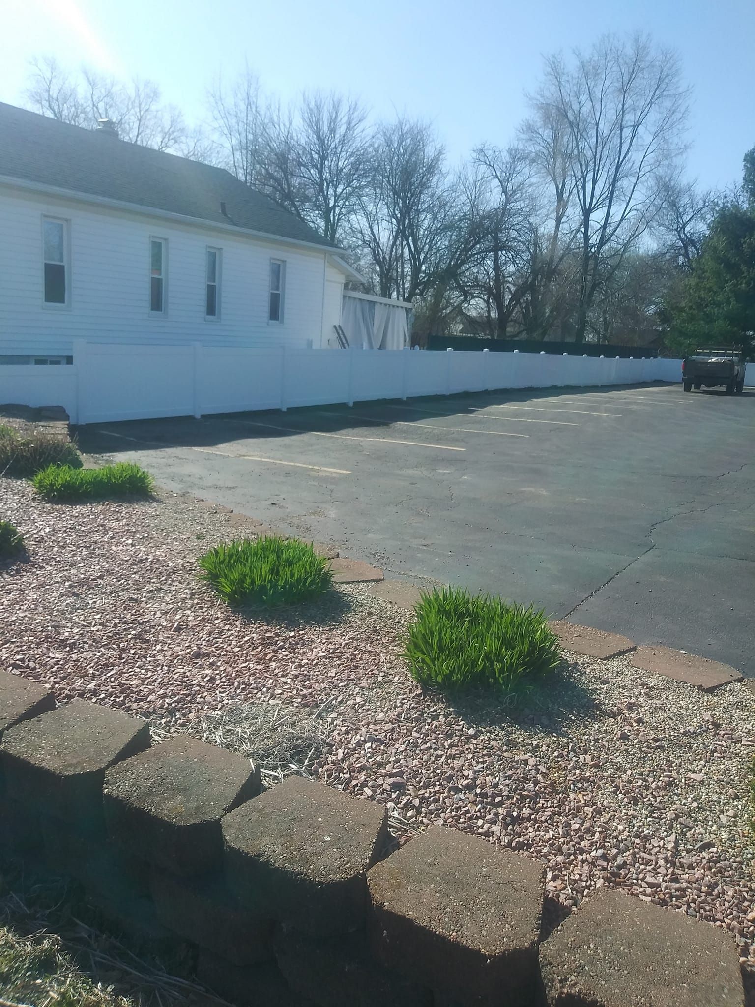 A white building with a privacy fence sits behind a gravel landscape border and an asphalt parking lot under a blue sky.