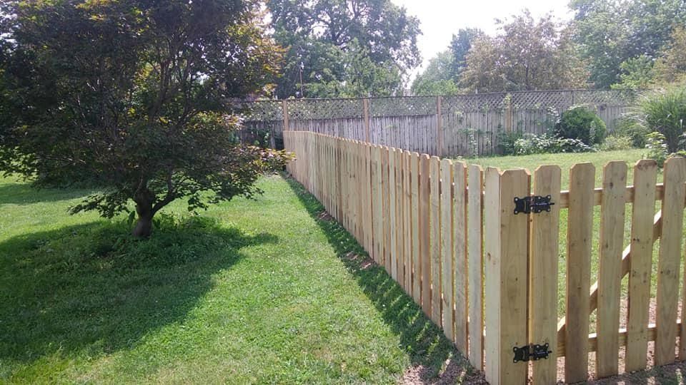 A wooden picket fence in a grassy yard, with a tree on the left and a darker fence in the background.