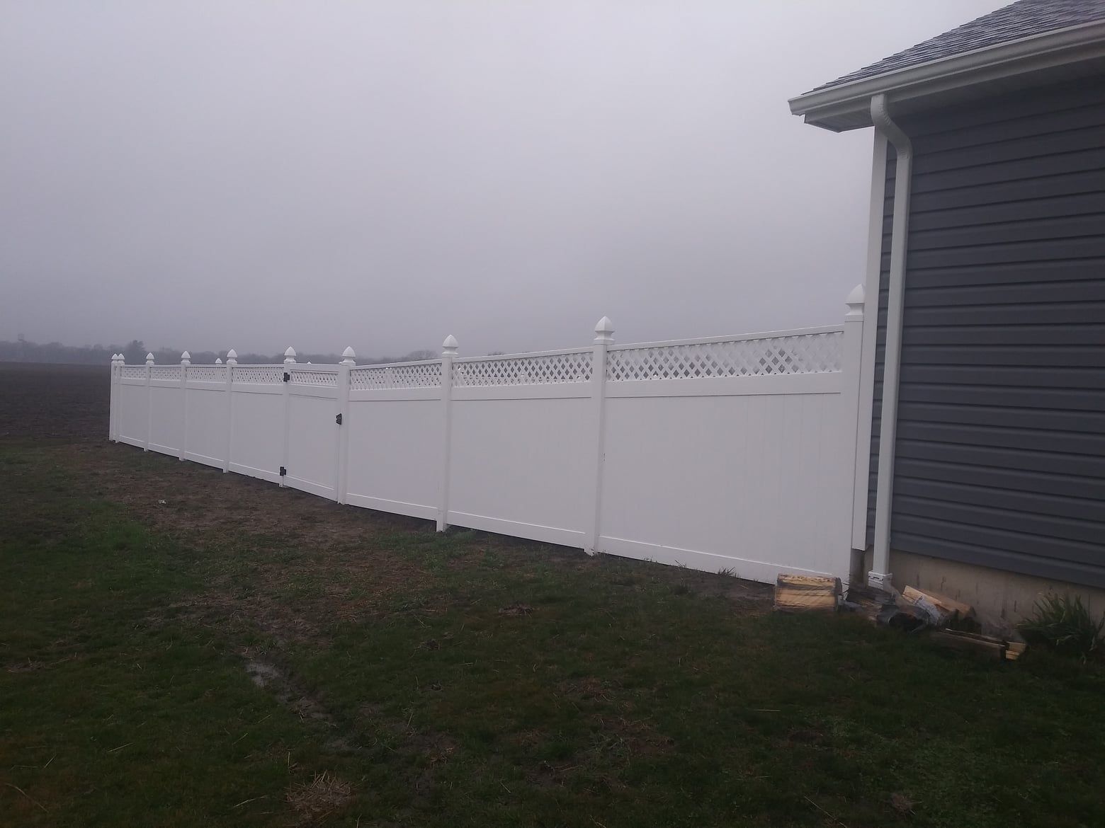 A white vinyl privacy fence with a lattice top extends from the corner of a grey house into a grassy yard under foggy skies.