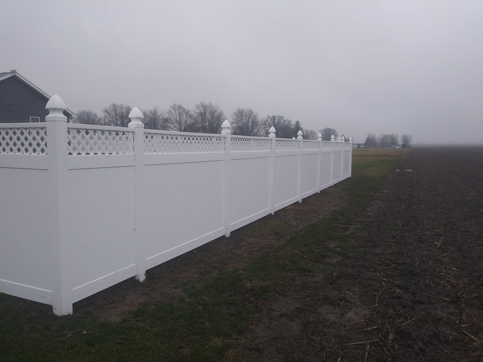 A long white vinyl fence with decorative lattice top, extending alongside a dark, plowed field under a gray, foggy sky.