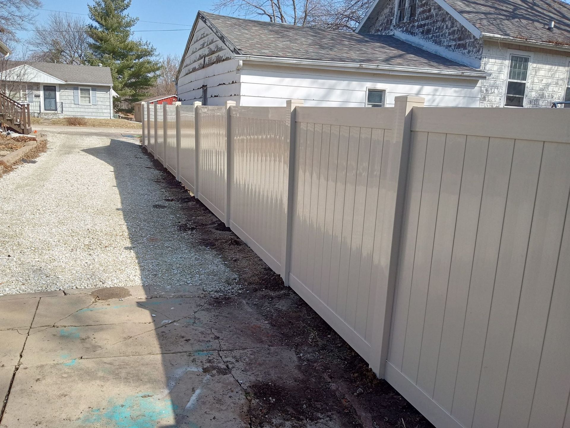 A beige vinyl privacy fence runs alongside a gravel driveway next to a residential house.